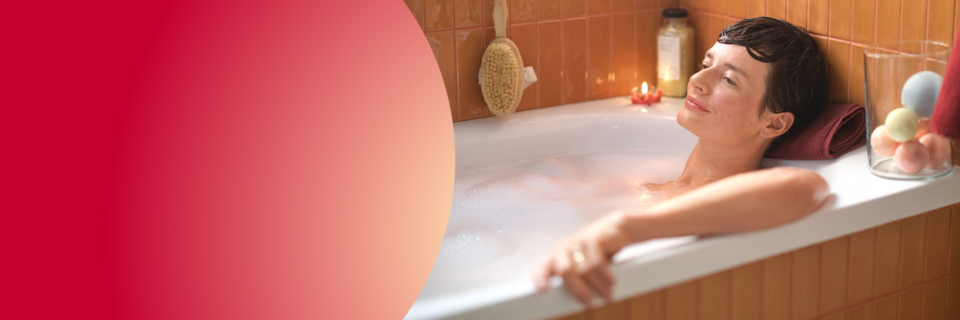 Kobieta relaksująca się w wannie pełnej piany, obok szklane naczynie z kulami do kąpieli i kosmetyki na brzegu – odkryj produkty do pielęgnacji ciała w Rossmann i znajdź chwilę dla siebie.